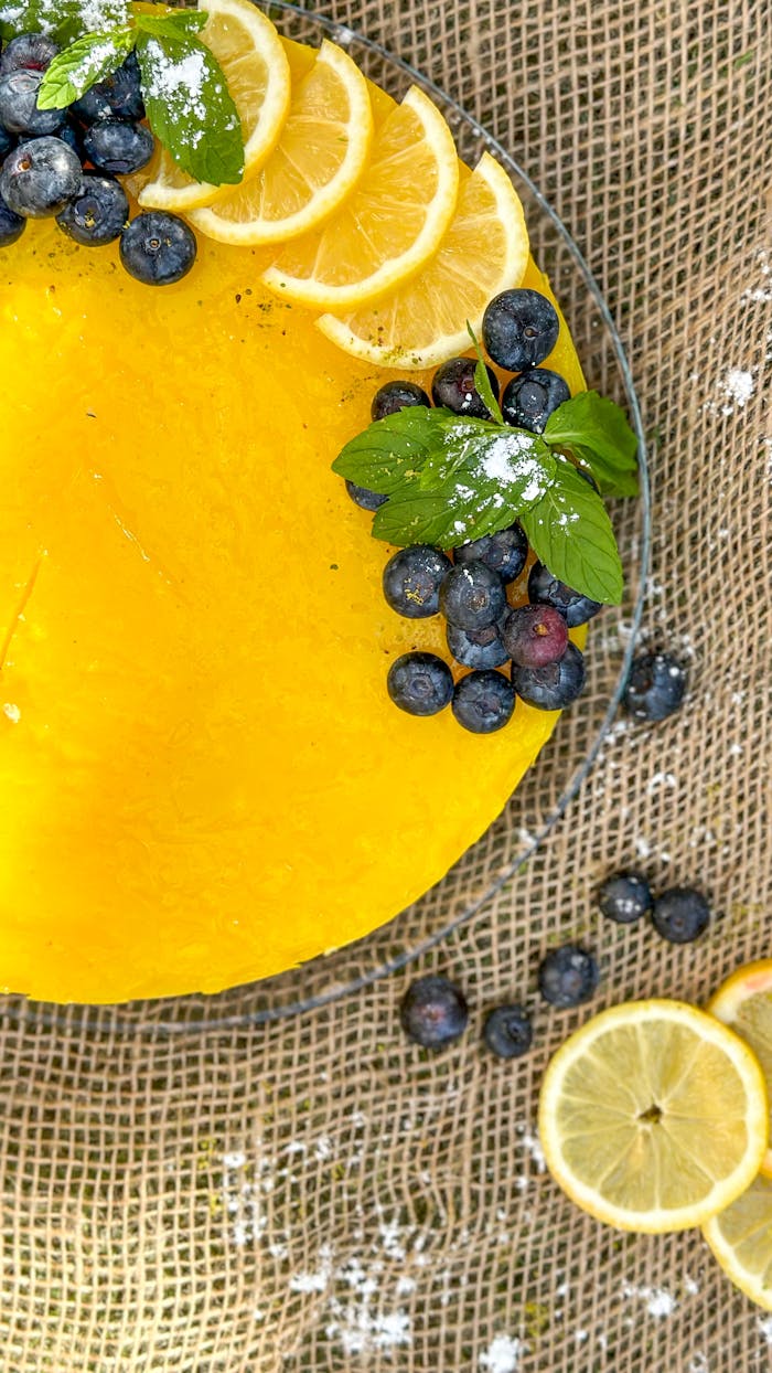 Colorful lemon cake topped with blueberries and mint leaves on a textured surface.