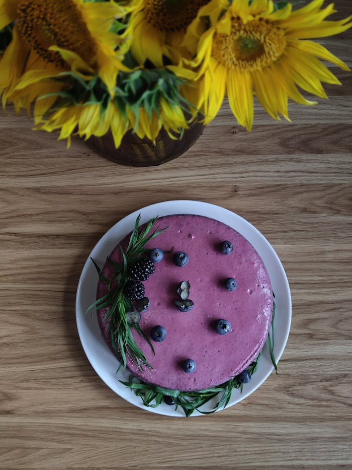 A purple berry cake garnished with blueberries on a wooden table, accompanied by sunflowers.