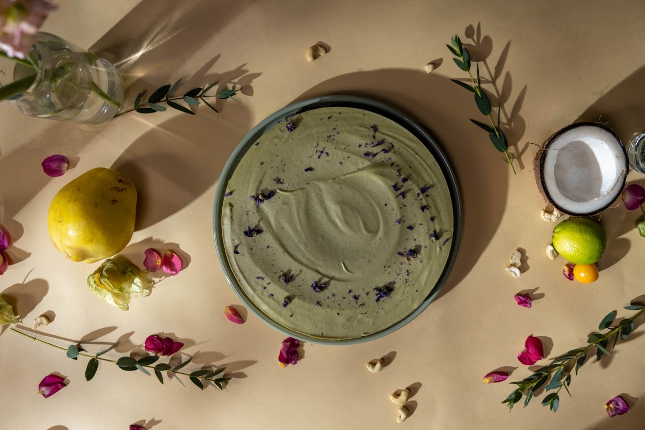 Top-down view of a vegan green cake garnished with flower petals, surrounded by fruits and leaves.